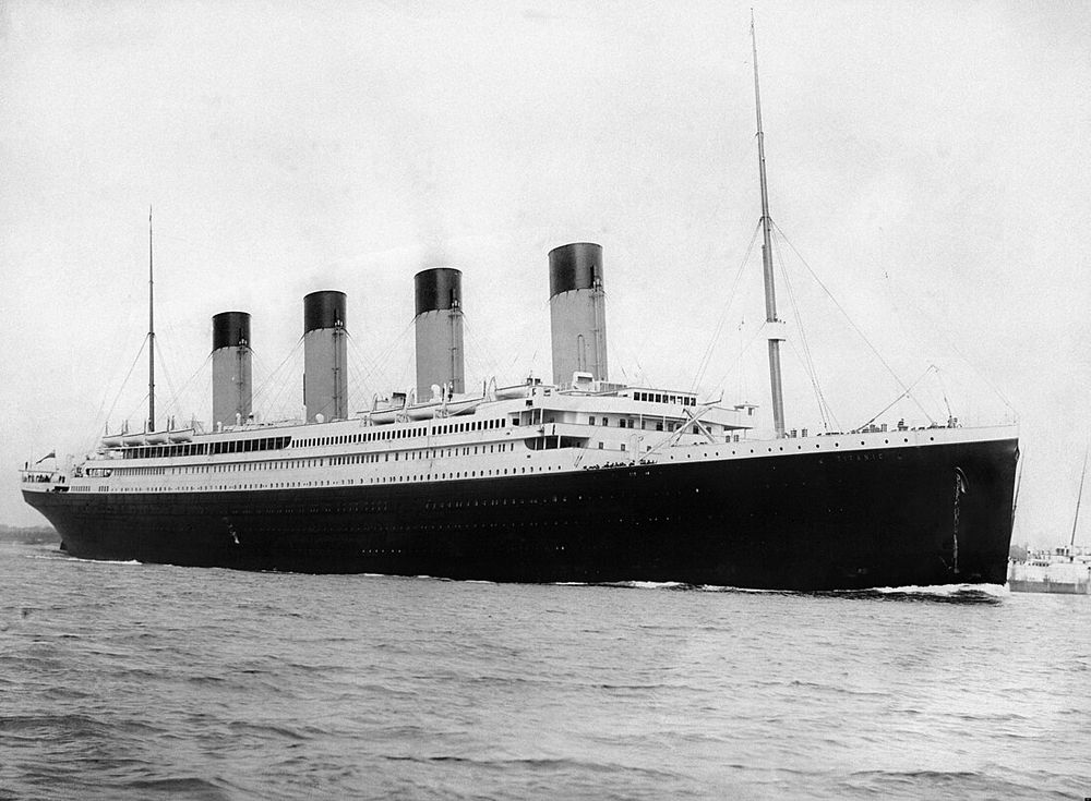 Schwarz-weiss-Fotografie des Passagierschiffs RMS Titanic auf dem Wasser, aufgenommen von der Seite; das Schiff hat vier markante Schornsteine und ist teilweise von Takelwerk und Masten umgeben.