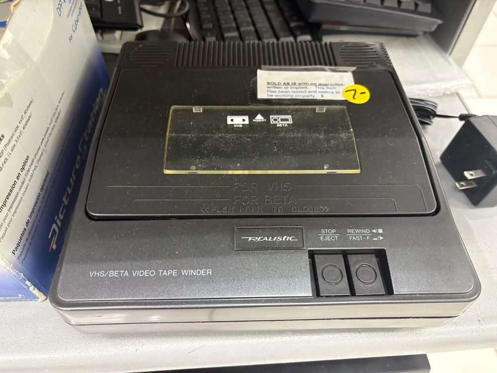 A VHS/Betamax tape rewinder sitting on the shelf at a thrift store. A round yellow sticker on it has the number 7 written in marker. There’s also a sticker with a bunch of text to the effect that it has been tested and seems to work but they are providing no warranty.