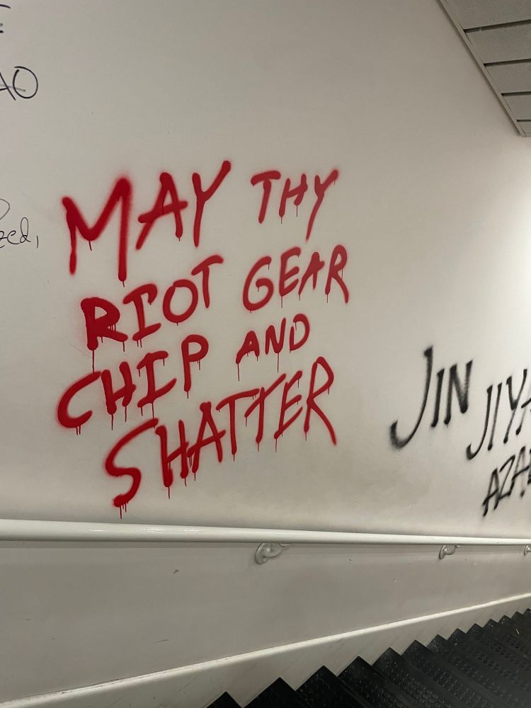 Tagged stairwell of Portland Stat University's Refaat Alareer library in Spring of 2024. The text of the tag is in a dripping red paint, over an off-white wall. The text reads "May thy riot gear chip and shatter"