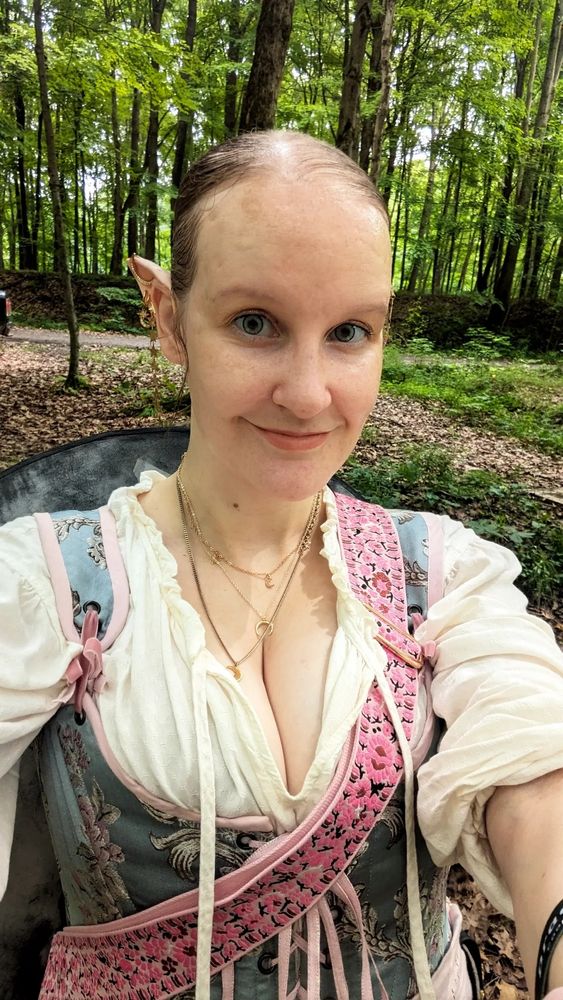 A selfie of a cute elven woman with pointy ears bedecked in moon-themed jewelry. She's dressed in blue stays over a white blouse, a shield strung over her shoulder on a pink floral strap.