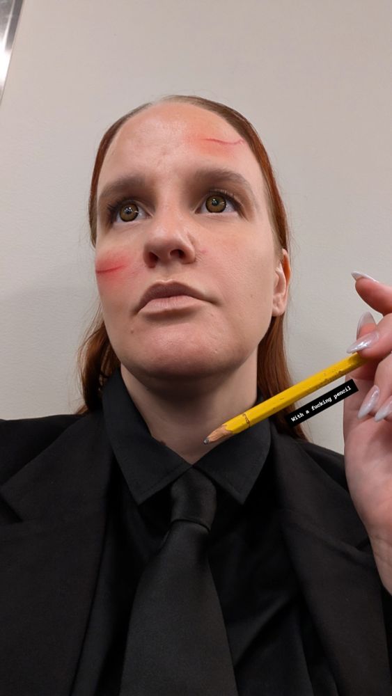 A woman dressed as John Wick in a black suit and tie holding a pencil. She appears to have fresh cuts on her face, but they are makeup. The text "with a fucking pencil" is present in the photo as well.