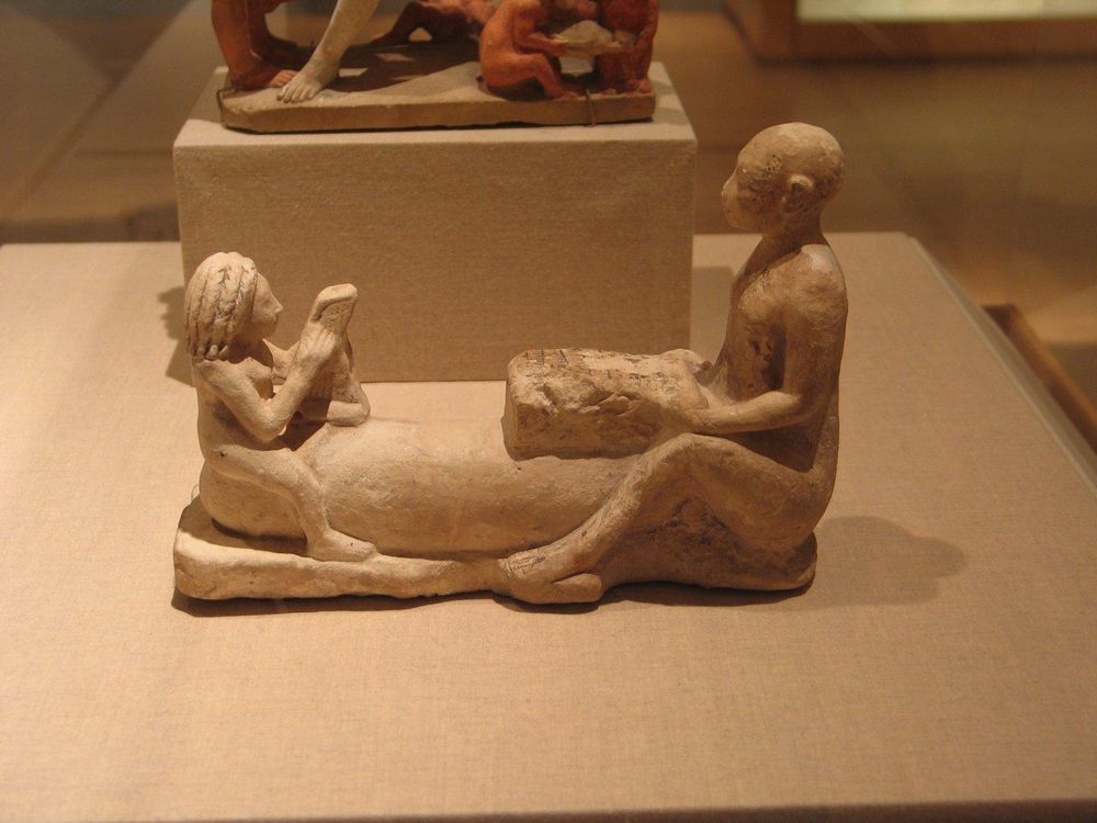 Figure group in soft white limestone of a woman and a man with an oversized phallus, dated 305-330 BCE. The nude man is seated, his enlarged phallus extended on ground. A smaller figure of a nude woman playing her harp is seated on the far end of his phallus. The man holds a curious object in front of him – a musical instrument? Some detail are outlined in black paint.