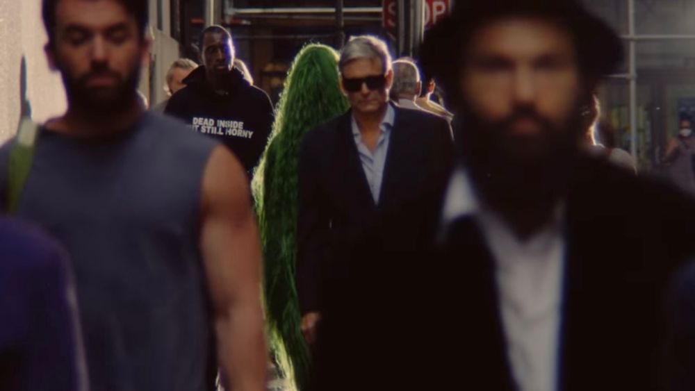 Another screenshot from “The Subway” MV showing a crowded street. A man just to the left of the woman with long green hair is wearing a black shirt that says “Dead inside but still horny”