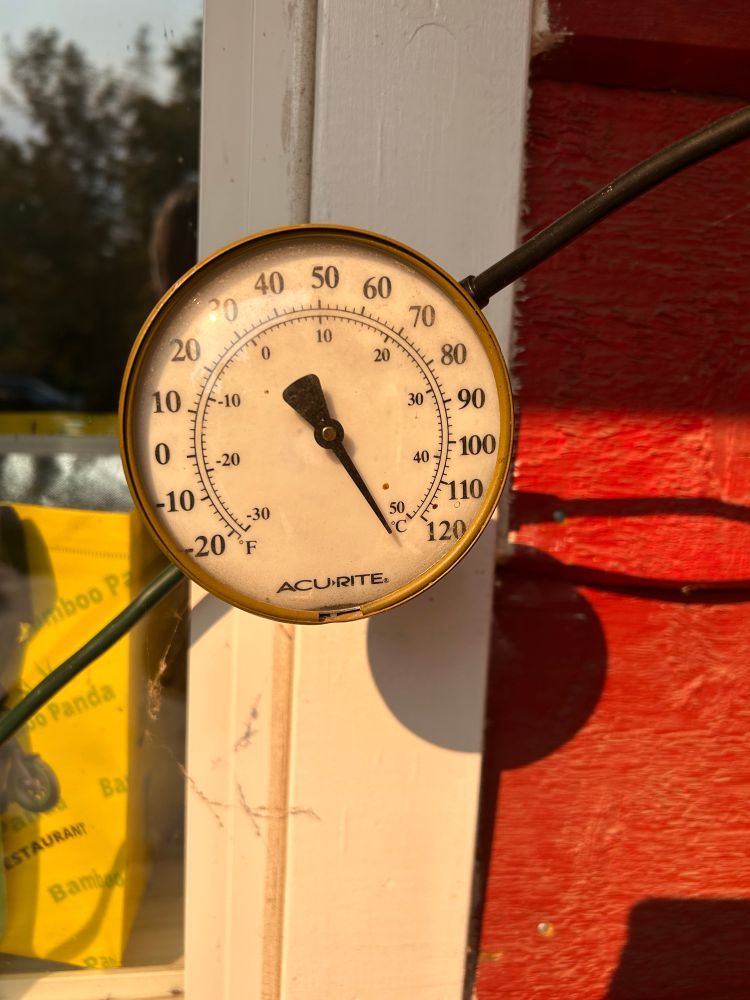 A very old Acu-rite thermometer reads past the 50c/120f marker as the sun shines down on it. It is attached to a windowsill a foot away from the front door of a house.