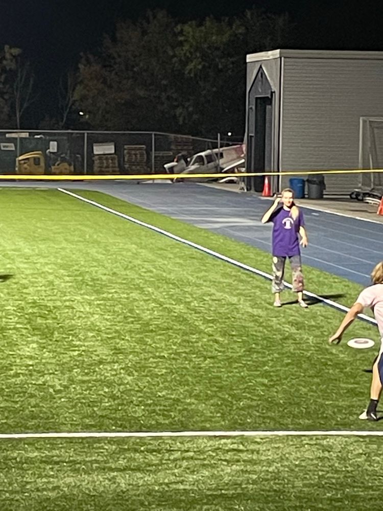 Brightly lit green pitch with frisbee throwers in foreground, small crashed plane in background.
