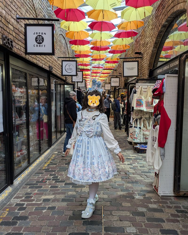 A person wearing a blue and white Alice themed bunny outfit, posing in the middle of an alley adorned with red, yellow, and orange umbrella above.