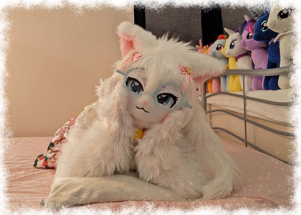 A white cat fursuiter laying on a bed and resting her head on both paws.