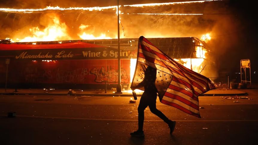A protester carries a US flag upside down, a sign of distress, past a burning supermarket.