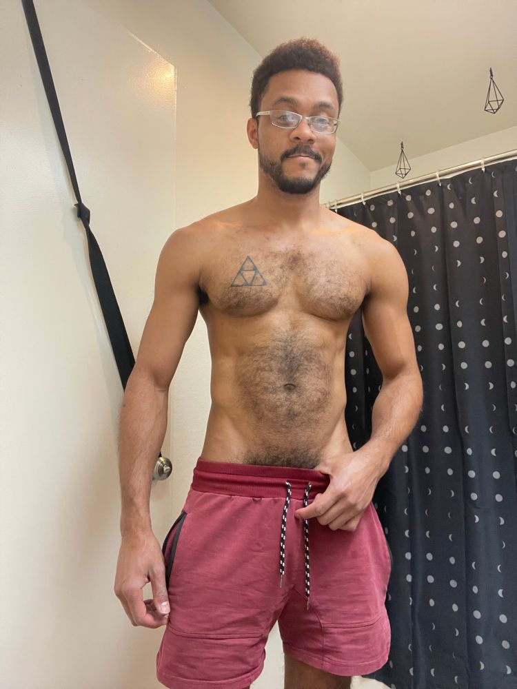 A black man standing in his bathroom; he is only wearing short red shorts, revealing torso lightly covered in curly black hair and a triforce on his pectoral