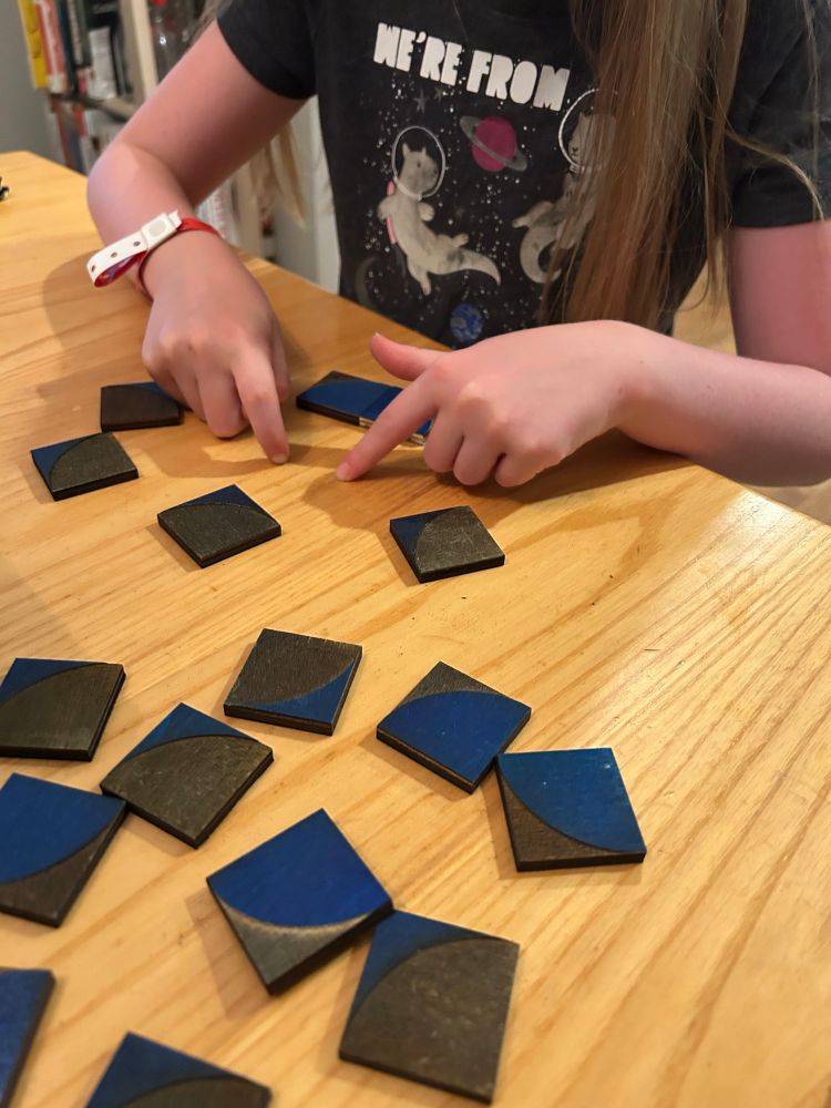 Several wooden square tiles laid out on a table. The square tiles have quarter circle segments and the arcs left over from a quarter circle segment on the.  Tiny hands are showing how they want the design to go. 