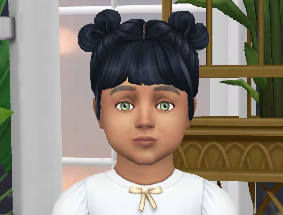 A Sims baby with black hair tied up in two buns. She has green eyes and is wearing a white shirt with a golden bow.