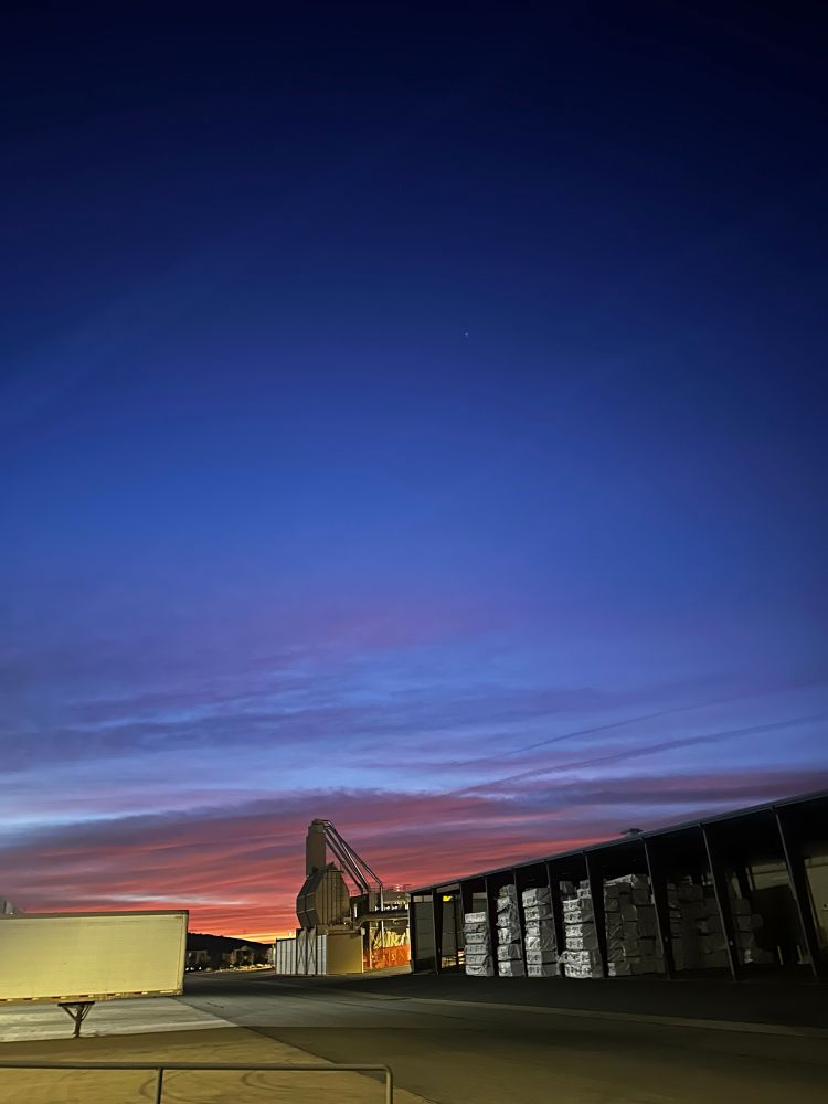 First sign of light at the warehouse. There is a mixture of pinks and purples in the clouds. A sawdust hopper and empty trailer are lit up by the warehouse lights. 