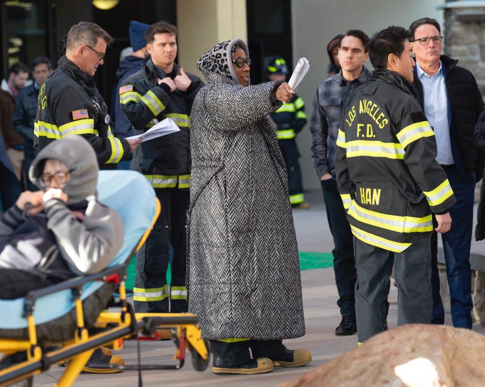 Aisha Hinds in a large coat which is on top of her firefighter uniform. She’s holding a rolled up piece of paper and is pointing with it. She’s directing an episode of 9-1-1 and her co-stars are around around her in their firefighter uniforms.