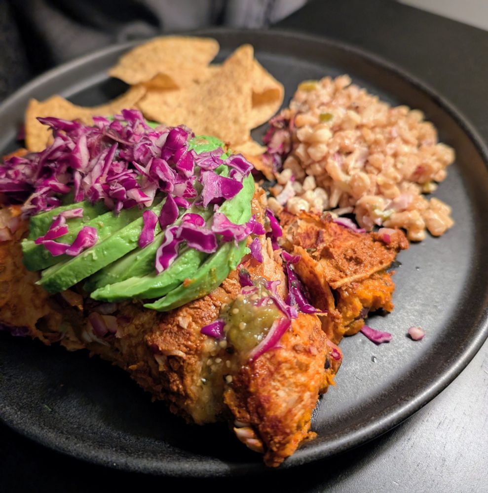 Three sisters enchiladas topped with avocado and purple cabbage, esquites and corn chips on a black ceramic plate on a black ground