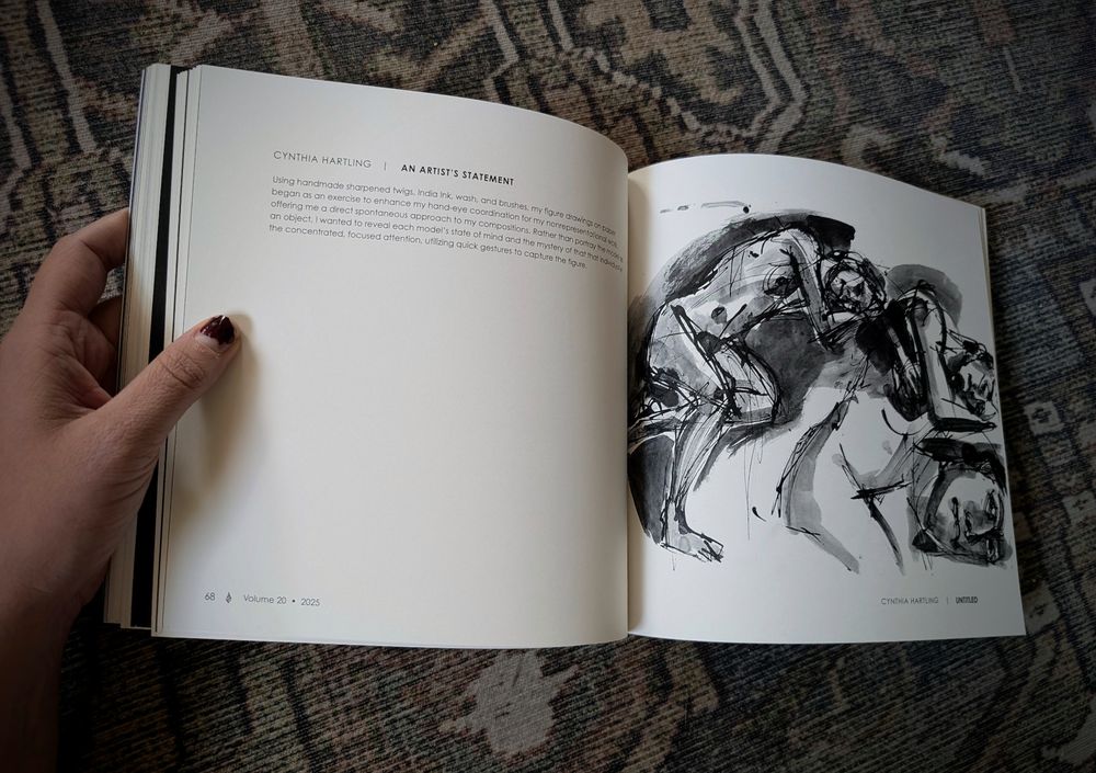 A hand opening a book on a green carpet, showing a drawing of three naked people lying on one another. 