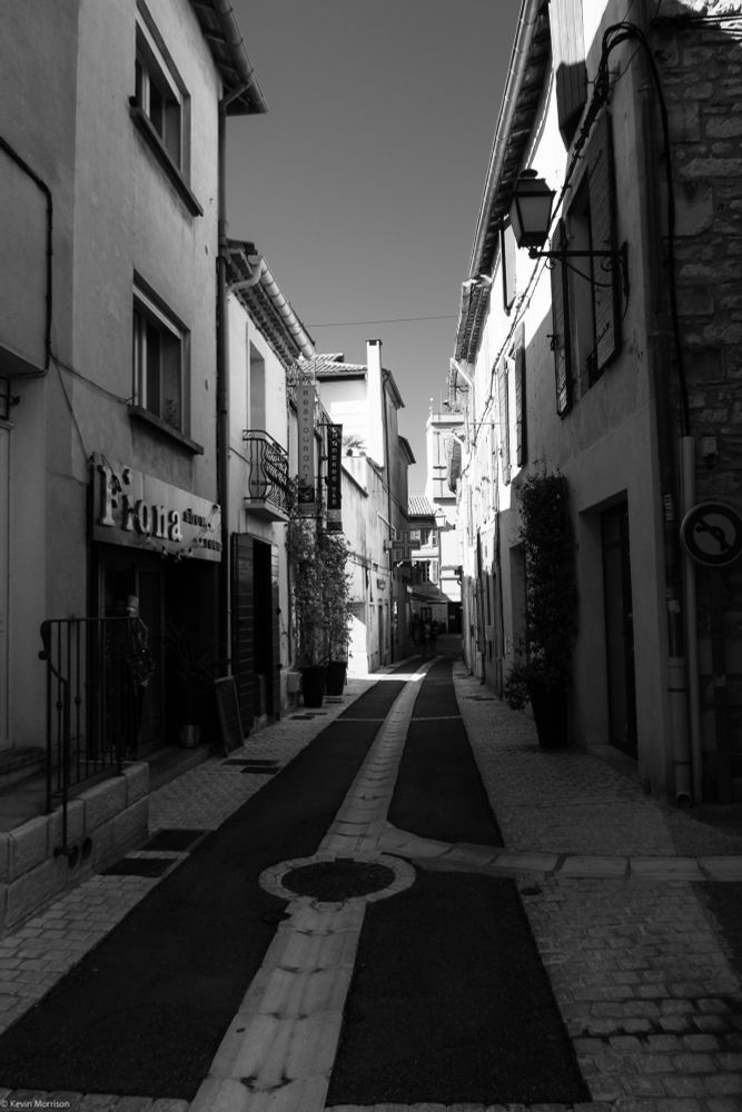 A narrow street in France with shops and homes on both sides, in shadow, brick pavement. black and white image. 