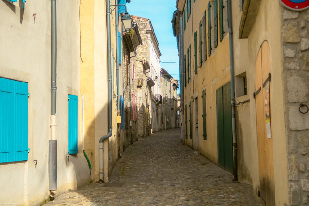 A narrow city street with buildings on both sides. Pastel color tones, cobblestone street. 