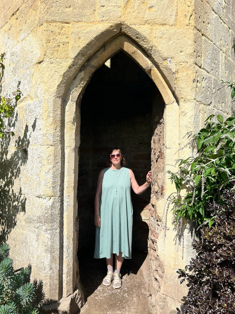 Helen in a pale green dress standing in the shade of a gothic arch outdoors 