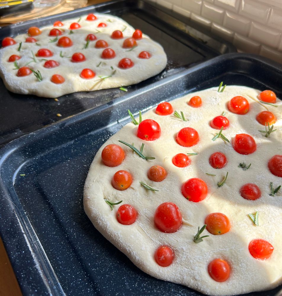 Two uncooked focaccia breads studded with cherry tomatoes and rosemary, rising in the sunshine 