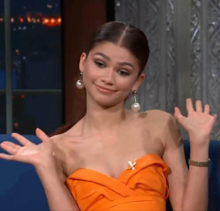 Zendaya in a orange top sitting down with her hands up 