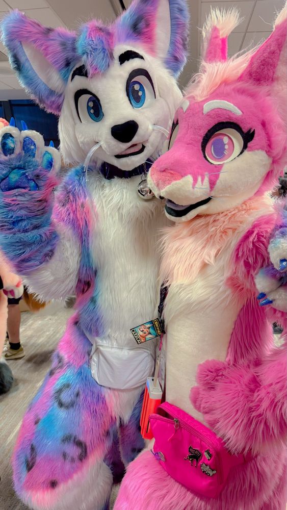 A white and galaxy kitty fursuiter (Astra) with a hot pink caracal fursuiter (Khora) posing together for a photo. Astra is waving and Khora has her hand on her hip