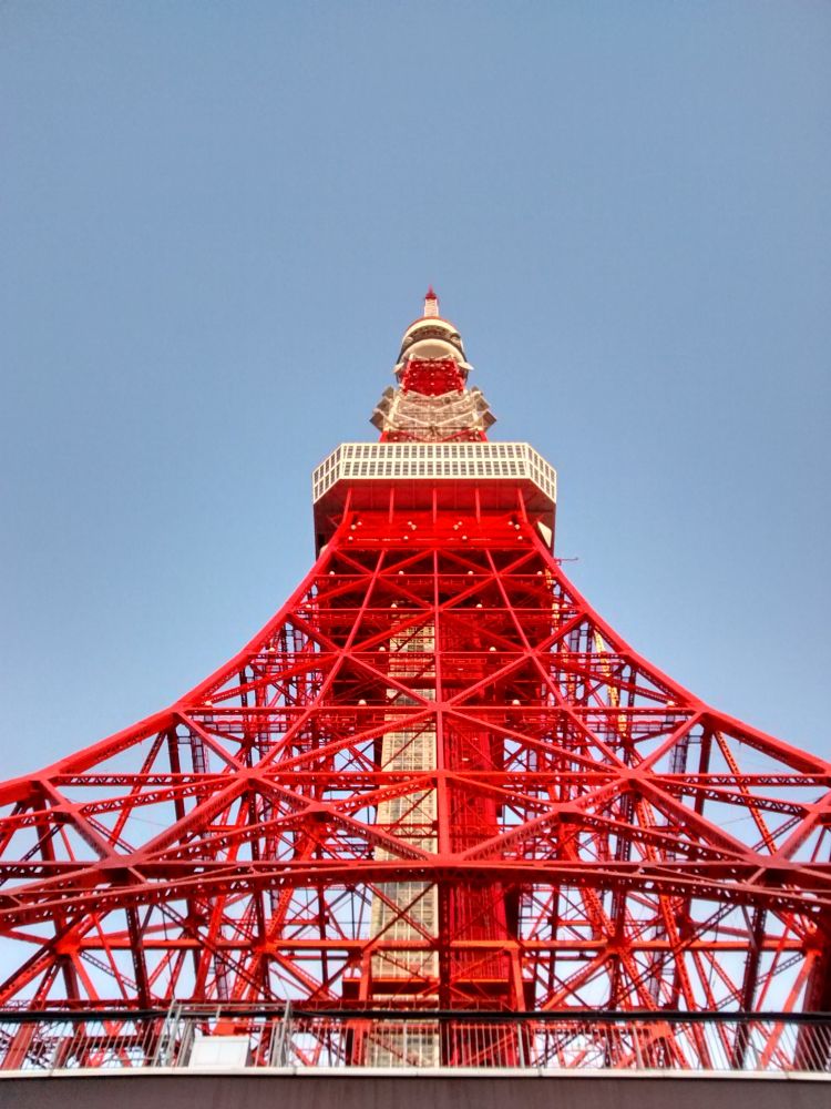 A photo of the red and white Tokyo Tower taken very close to the base, looking up. The latticed red metal structure, similar in structure to the Eiffel Tower, ascends to a point at the top. There is a large white viewing platform with a red base about two thirds of the way up, and a smaller round viewing platform much closer to the top. Through the middle of the structure, two tall pillars in red and white. Blue sky