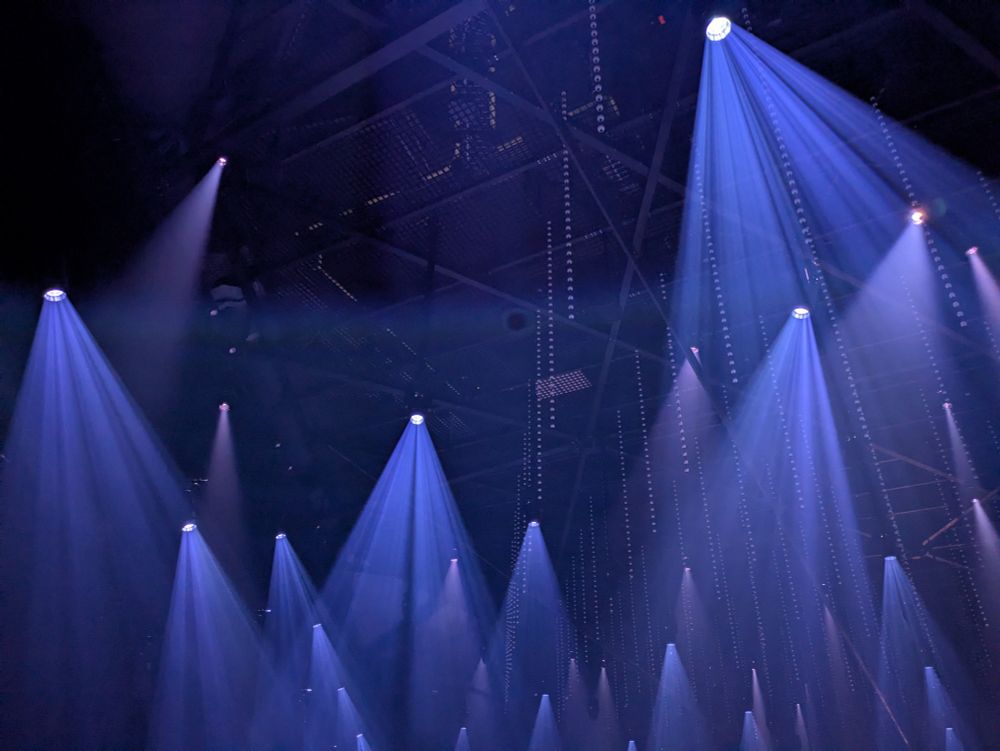 Blue spotlights at a music venue. Each light projects a blue cone of light downwards. Strings of LED ball lights can also be seen hanging from the ceiling