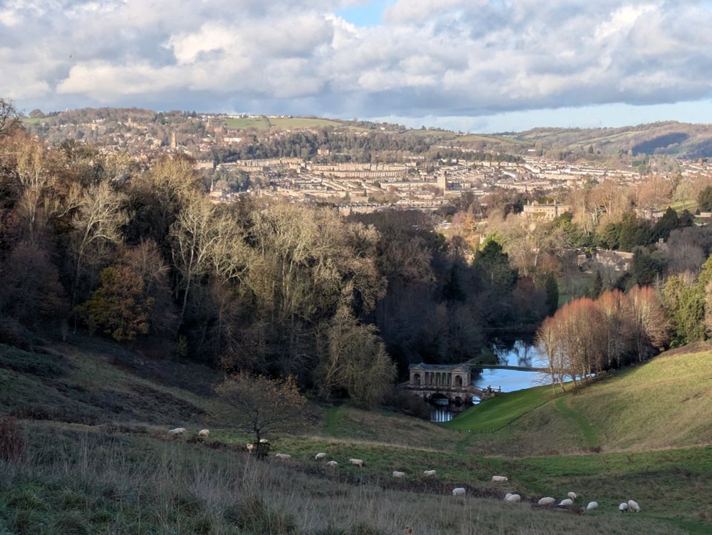 Photo taken from the top end of Prior Park down towards Bath. Its Georgian buildings can be seen in the distance. In the foreground green hillside drops away, with a flock of sheep grazing. Further down, a Palladian bridge crosses a small lake. Trees surround the park. On the other side of the city, hills are rising in the distance. Overcast sky with patches of blue