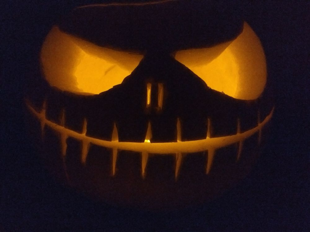 The spooky, illuminated face of a carved pumpkin with big eyes, two small slits for a nose and a wide thin grin with regular stitches. The photo is taken in darkness , little of the external colour of the pumpkin is visible. The internal candle illuminates all of the carved features in an orange glow 