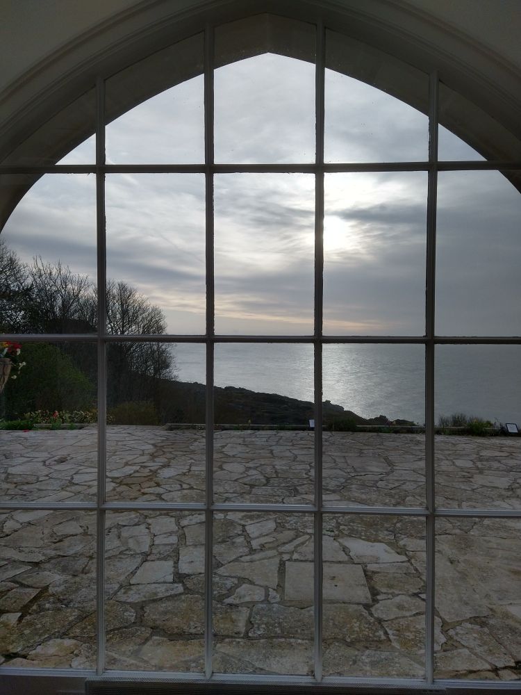 An arched window frame divided into 20 smaller mostly rectangular panes (apart from at the top at the arch looking out onto an empty crazy-paved stone patio.  Beyond the patio the land drops away and a calm sea can be seen, reflecting the morning sunlight.  The sun is nearly breaking through the mostly overcast sky. To the left of the frame, beyond the patio, trees.