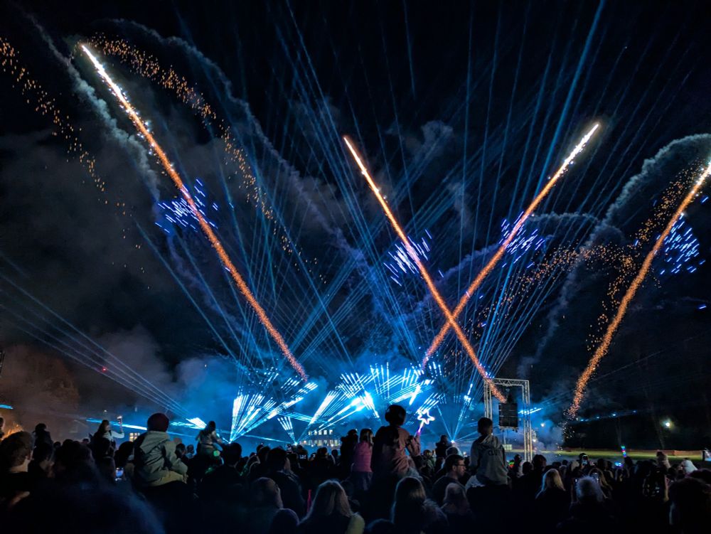 In the UK, fireworks are generally reserved for New Year's Eve and Bonfire Night in November. Here, orange, blue and white fireworks are going off amongst a blue laser show. In the foreground, a crowd watching the fireworks, some children sat on a parent's shoulders