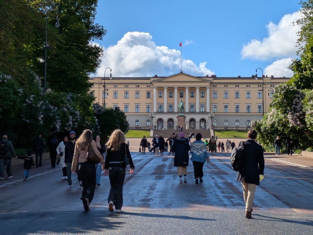Approaching the royal palace in Oslo on foot from Karl Johan's gate. In the foreground, a number of people are walking up and down the road leading to the palace. The road is mainly in shade from the trees alongside the road.  Further in the distance, in the sunshine, the yellow painted palace building is framed on either side by the trees, its central part with 6 columns and arches beneath stand behind a statue of King Karl Johan on horseback. A blue sky with fluffy white and grey clouds can be seen above the palace