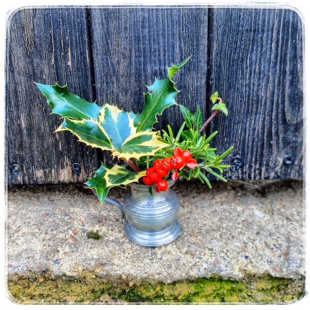 A mini (1/4 gill measure if you fancy knowing) pewter jug perched on a stone garden step with a weatherworn black painted wooden gate behind, the jug holding sprigs of green leaves foliage & bright red berries.