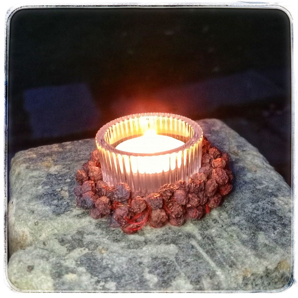 A tea light candle in a small glass holder with a ring of very dried out rowan berries set about it, sat on a stone gatepost in a darkening garden.