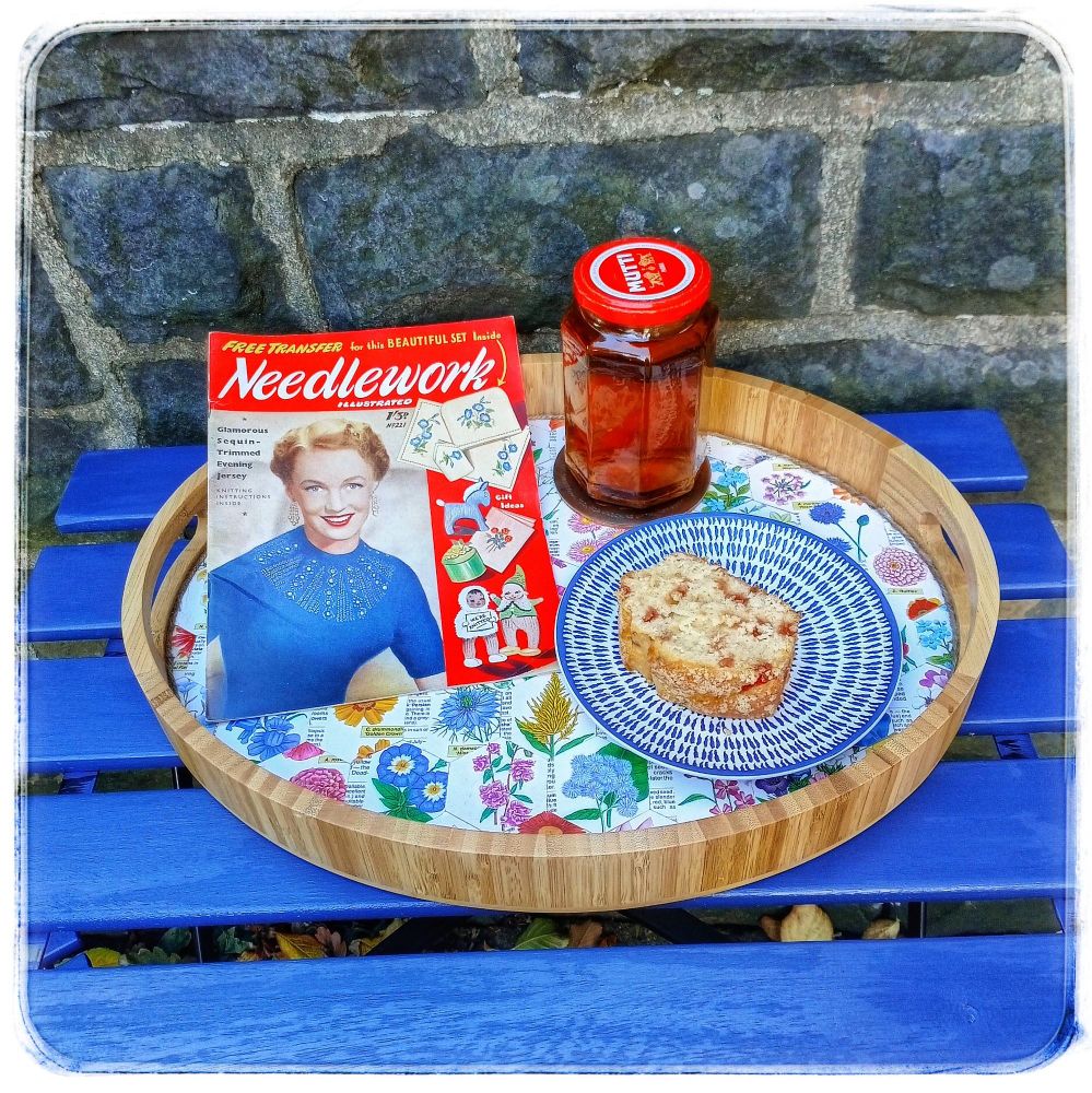 Against a stone wall is a quite blue painted wooden garden table with a floral decorated tray sat on it, bearing cake on a blue china plate, a jam jar of tea, & a 1950s magazine of handicrafts.