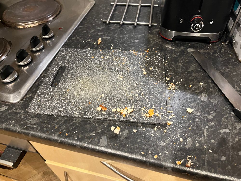 A countertop with an empty bread board and a bread knife. Crumbs of the bread have been scattered everywhere. 