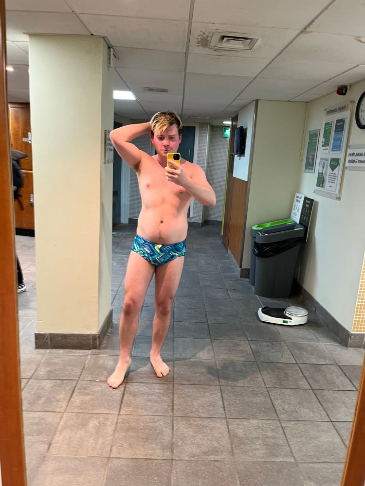 I stand in front of a gym changing room mirror taking a selfie. One hand is behind head. I wear short green-blue swimming trunks