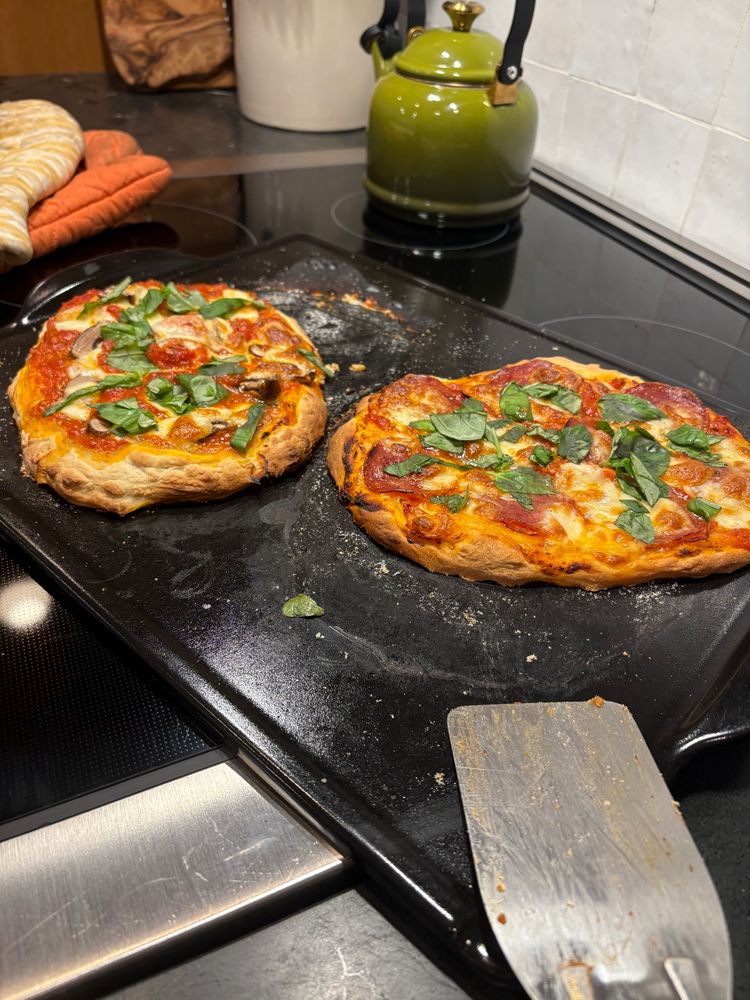 two homemade pizzas on a large black bread-baking enameled platter in front of a green tea kettle