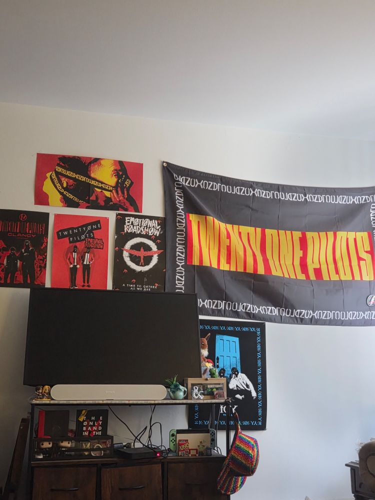 Various twenty one pilots posters and a flag are pinned to a wall behind a tv stand. There are two from emotional roadshow, one from the Clancy listening experience, the clancy album insert poster, a black velvet one from icy tour, and a flag from the Clancy tour. There is also a selection of memorabilia arranged on the TV stand itself, including confetti from every twenty one pilots concert the poster has been to and the funko pops of the band members. 

Some of the items are related to other interests of the post maker including D&D, Tangled, and Pokémon. A glittery rainbow cowboy hat hangs from the side of the TV stand. 