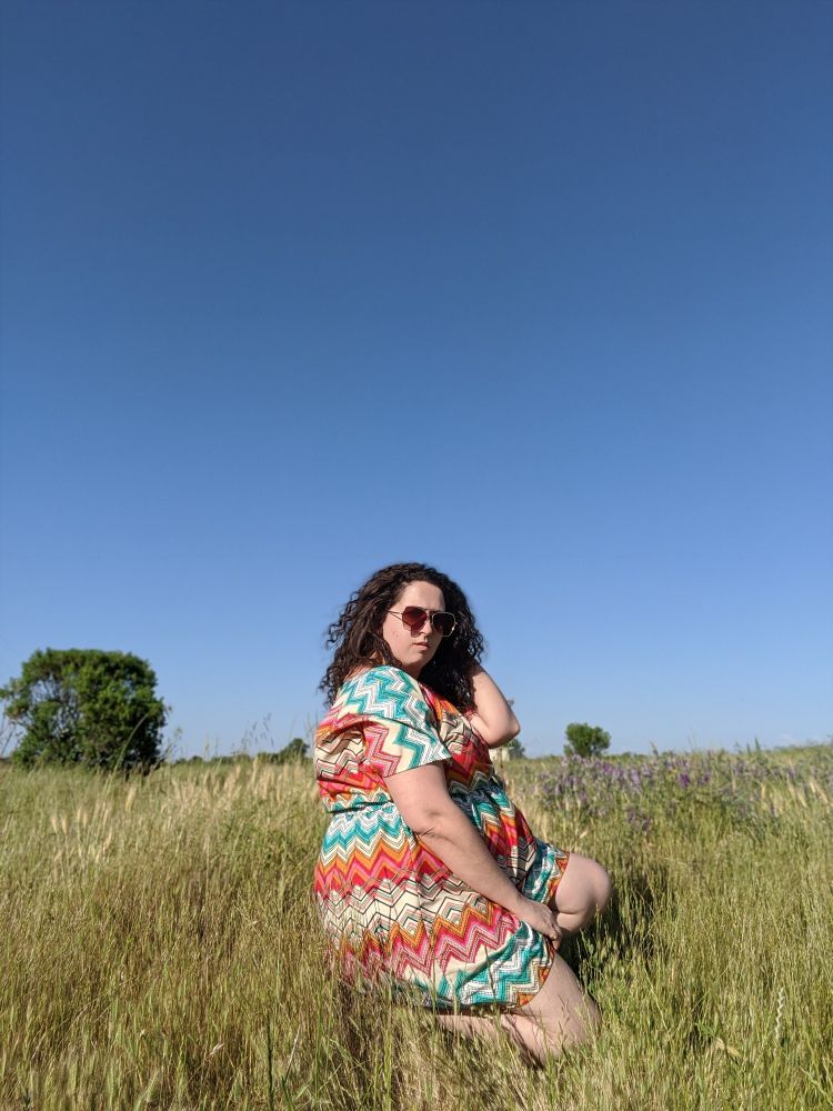 White femme presenting person crouching in field with clear blue sky behind them. They are wearing a multicolored dress and sunglasses.