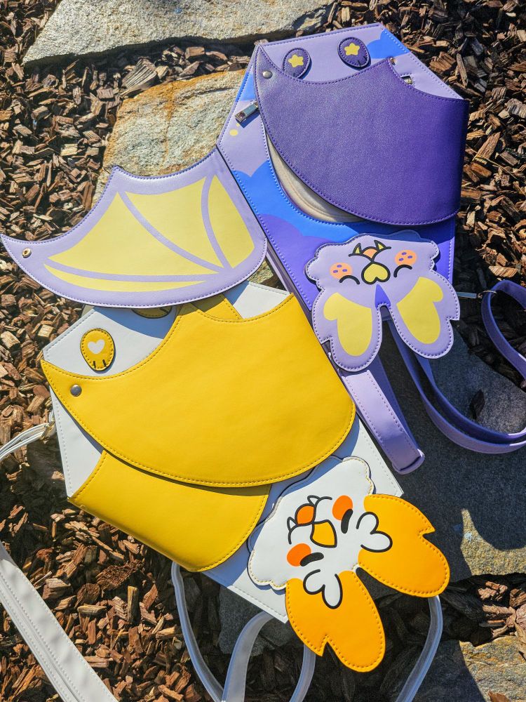 A photo showing two bat shaped backpacks, or "batpacks", one colored like a honduran white bat & the other colored in dreamy yellows & purples.