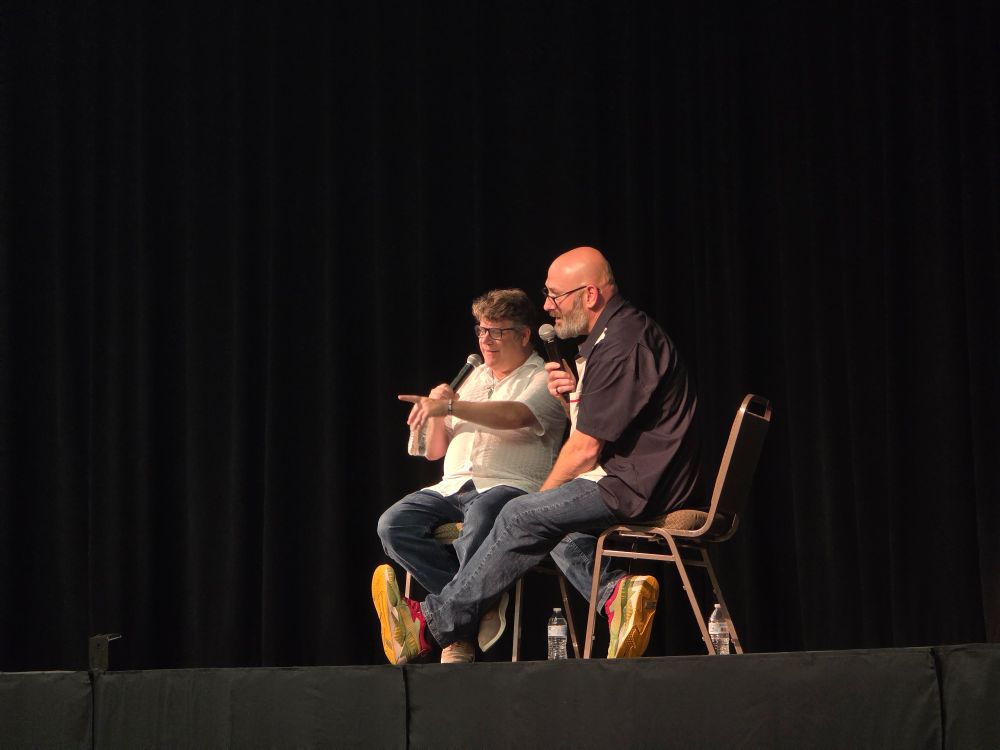 two men sit on a stage and point toward an audience