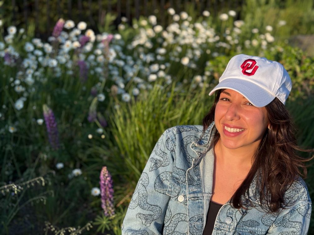 Anat (me) wearing a white hat with red OU logo, jean jacket with a snake design, and a huge smile! Lupines and daises are in the background.