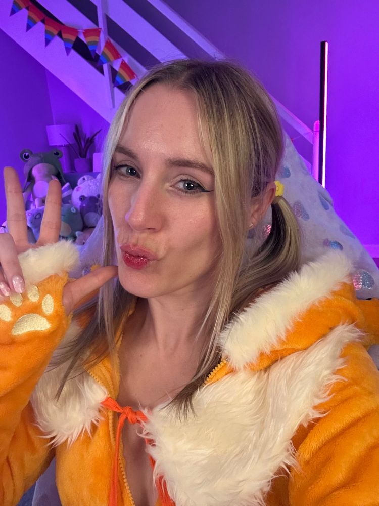 A selfie of Melzelia. She's in an orange fur outfit, with cream coloured fur trim around the hood and hands. It has cream coloured paw prints on the hands of the long sleeves. There is a cream fur shawl around her shoulders, tied with an orange string at the front. Mel has pale skin and blond hair, which is pulled into pigtails with long bangs framing her face. She has blue eyes and pink lips. She is doing a kiss at the camera, with one hand up near her face in a peace sign. In the background are lots of frog plushes, lit up in purple lighting.