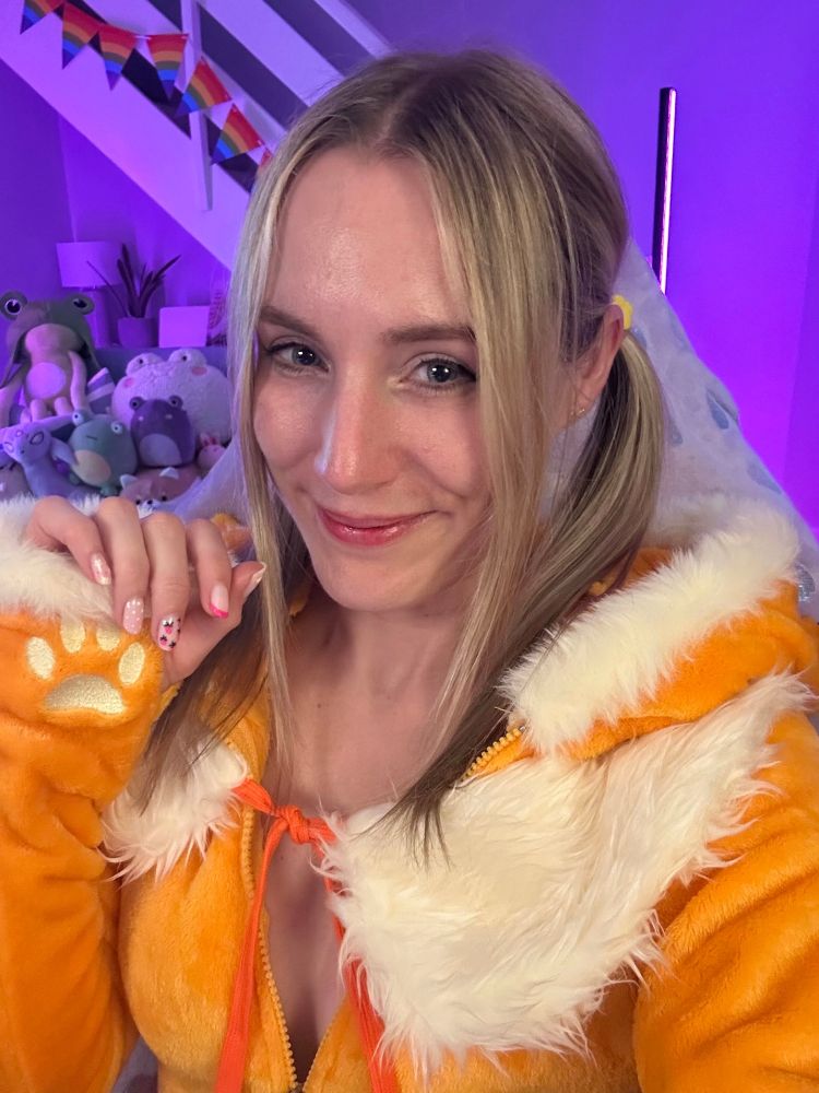 A selfie of Melzelia. She's in an orange fur outfit, with cream coloured fur trim around the hood and hands. It has cream coloured paw prints on the hands of the long sleeves. There is a cream fur shawl around her shoulders, tied with an orange string at the front. Mel has pale skin and blond hair, which is pulled into pigtails with long bangs framing her face. She has blue eyes and pink lips. She is smiling at the camera, with one hand up near her face, kind of like a paw. In the background are lots of frog plushes, lit up in purple lighting.