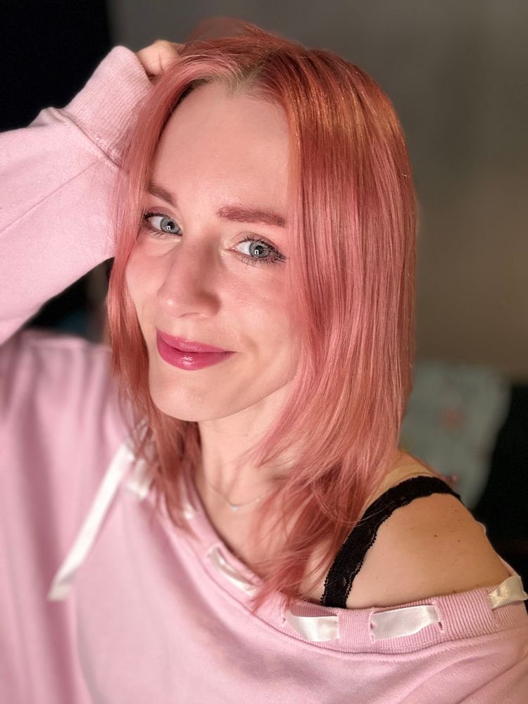 A selfie of Melzelia who has pale skin, blue eyes, pink lips, and rose pink hair. She is smiling at the camera and has one hand in her hair. She wears a long sleeve pink sweatshirt with white ribbon woven through the neckline, revealing one shoulder and a black lacy strap.
