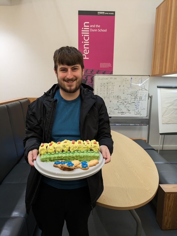 Matthew Clark holding a tray with a layered rectangular cake, showing the periodic repeating structure of IFT-A and IFT-B. Gingerbread cookies cut in the shape of dynein-2 ride on top.