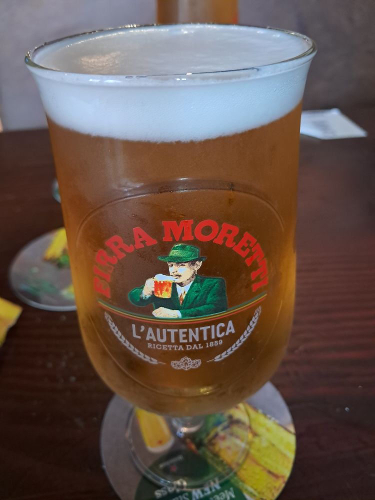 A pint of Birra Moretti. Golden beer, white head, on a brown table. The logo and a man drinking the beer on the front. 
