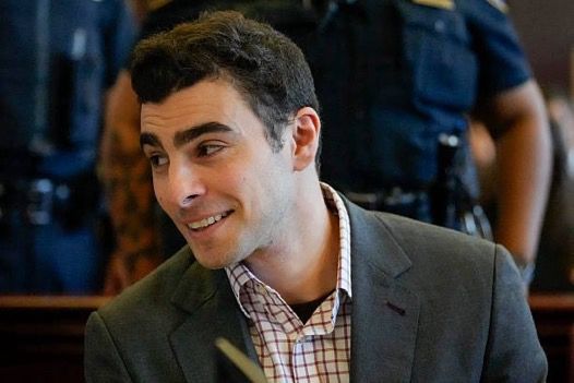 Luigi Mangione olhando de lado, com as sobrancelhas arqueadas, a testa fazendo ondinhas, e ele sorri. Ele veste paletó cinza, camisa xadrez branco com listras vermelhas, e camiseta por baixo da camisa. 