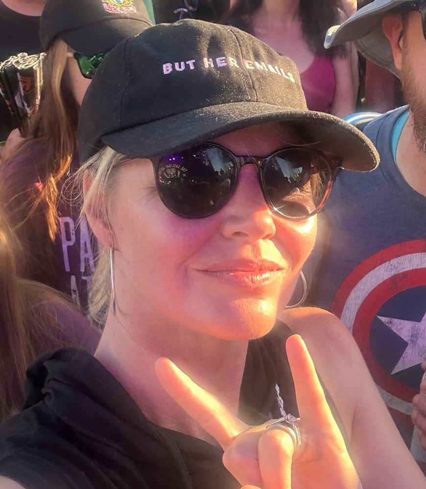 White lady in sunglasses, sun reflecting off her face, giving with two fingers up and wearing a black hat with pink writing that says “but her emails.”